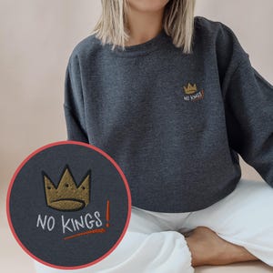 Könnte beinhalten: Ein dunkelgraues Sweatshirt mit Rundhalsausschnitt und einer Stickerei auf der linken Brust. Das Motiv zeigt eine goldene Krone über dem weißen Schriftzug "NO KINGS", ergänzt durch ein orangefarbenes Ausrufezeichen und einen orangefarbenen Strich.