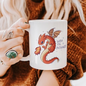 Op de afbeelding: Een witte keramische mok met een cartoonillustratie van een kleine rode draak die vuur spuugt. De tekst "burn the PATRIARCHY" staat in het blauw naast de draak gedrukt. De mok heeft een gladde afwerking en een standaard handvat.