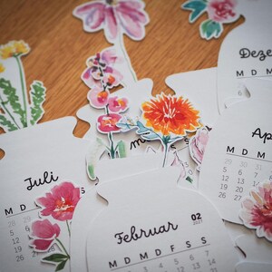 Verschiedene Kalenderblätter mit Aquarell-Blumenmotiven und Monatsnamen wie Februar, Juli, April und Dezember auf Holzuntergrund verstreut.