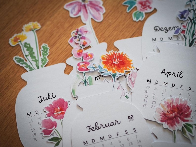 Verschiedene Kalenderblätter mit Aquarell-Blumenmotiven und Monatsnamen wie Februar, Juli, April und Dezember auf Holzuntergrund verstreut.