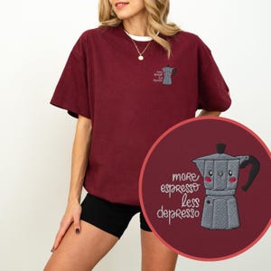 Puede incluir: Camiseta burdeos con cuello redondo blanco. La camiseta presenta una cafetera bordada con el texto "more espresso less depresso". Una versión más pequeña de la cafetera está bordada en la parte superior izquierda.