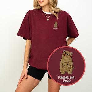 Könnte beinhalten: Ein kastanienbraunes T-Shirt mit kurzen Ärmeln und einer kleinen gestickten braunen Bärenfigur auf der linken Brust. Unter dem Bären steht der Text "I Choose the Bear" in Weiß. Ein kreisförmiger Ausschnitt zeigt eine vergrößerte Ansicht.