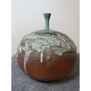 May include: A round ceramic vase with a narrow neck, featuring a smooth terracotta-coloured base and a textured upper section with a glaze that drips in shades of cream, brown, and light blue, creating a layered effect on the spherical body.