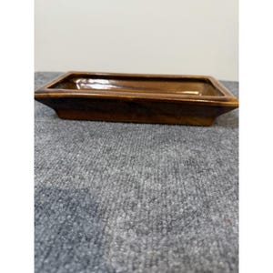 May include: A rectangular ceramic tray with a glossy brown glaze. The tray features a shallow basin with slightly angled sides and a flat rim, showcasing a smooth, reflective surface with subtle variations in the brown colour tones throughout.