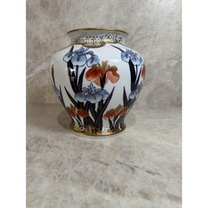 May include: A white ceramic vase featuring a hand-painted floral pattern of blue and orange irises with dark green foliage. The vase has a rounded body, a flared rim, and gold-toned trim along the top edge and base, with gold accents.