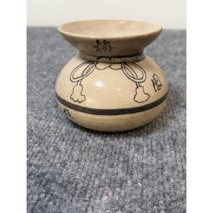 May include: A small, beige ceramic vessel with a wide, flared rim and a rounded body. The piece features hand-painted black line art depicting a decorative knot with tassels, along with black Japanese characters and a horizontal band encircling the base.