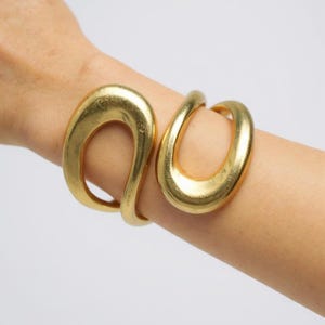 May include: A gold-colored cuff bracelet with an open, abstract design. The bracelet features two large, oval-shaped elements connected by a curved band. The metal has a slightly textured appearance, reflecting light.
