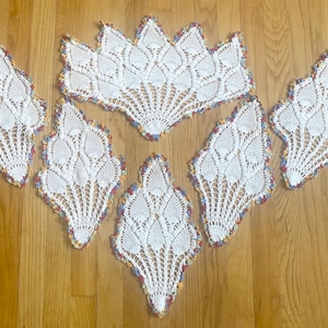 May include: A set of six white crocheted doilies featuring intricate pineapple-style patterns and scalloped edges trimmed with variegated pastel-colored yarn. The collection includes one large fan-shaped piece and five smaller diamond-shaped pieces.