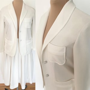 May include: A two-piece white suit set, consisting of a blazer and a skirt. The blazer has a notched lapel, two front pockets, and button closures. The skirt is pleated. The suit is displayed on a mannequin.