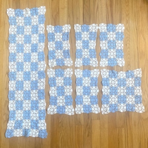 May include: A set of six crocheted doilies and a table runner featuring a checkered pattern of light blue and white squares. Each piece is crafted with an intricate floral-like openwork design and finished with a scalloped edge on all sides.