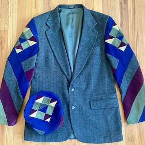May include: A gray blazer with patchwork sleeves in blue, purple, and green geometric patterns. A matching blue hat with a patchwork design is placed near the blazer. The blazer has a two-button closure and a light blue lining.