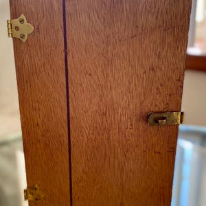 May include: A rectangular wooden storage case with a visible grain pattern, featuring a brass handle on the top, two decorative brass hinges on the left side, and a brass latch mechanism on the right side to secure the closed lid.