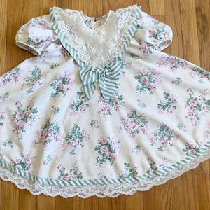 May include: A vintage white dress with a floral pattern of pink roses and green leaves. The dress features a lace collar, short puff sleeves, and a striped bow. The hemline is trimmed with lace and a green striped border.