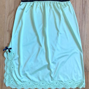 May include: A light green slip skirt with a black lace trim at the waist and a matching lace hem. A small navy blue bow is attached to the side. The skirt is made of a smooth, silky material.