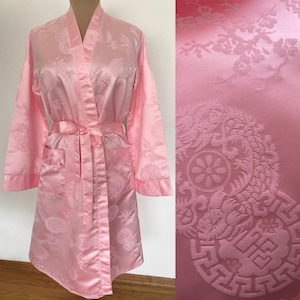 May include: A light pink satin robe with a matching belt and pockets. The robe features a subtle, repeating pattern of circular and floral designs. The fabric has a slight sheen, and the robe is displayed on a mannequin.
