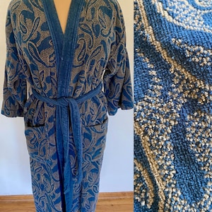 May include: A long-sleeved robe featuring a blue and tan paisley pattern. The garment includes a matching tie belt, two front pockets, and a shawl collar. The fabric has a textured, terry cloth-like appearance with a dense, looped pile.