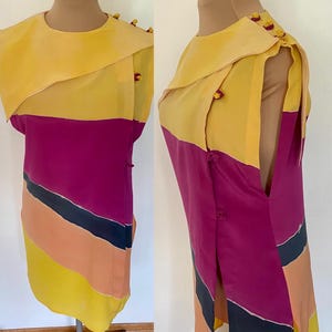 May include: A vintage, color-blocked dress in shades of yellow, fuchsia, black, and peach. The dress features a unique asymmetrical collar and button details on the shoulder and side. The dress is displayed on a mannequin.