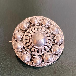 May include: A silver-toned brooch with a circular design. The brooch features a central, raised, round element surrounded by a ring of smaller, spherical accents. The outer edge is detailed with additional spherical embellishments, creating a textured, ornate appearance.