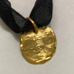 May include: A gold-colored pendant with a textured surface hangs from a black ribbon. The pendant has a design on the front.