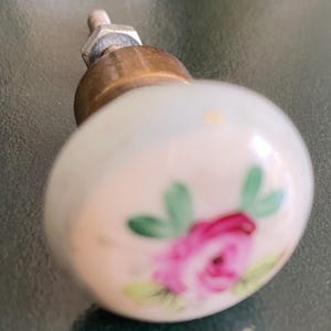 May include: A small, round, white ceramic cabinet knob with a floral design. The knob features a pink rose with green leaves. The base is brass-colored, and the screw is silver. This decorative knob is suitable for furniture.