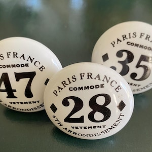 May include: Three white cabinet knobs with black text. Each knob is labeled "PARIS FRANCE COMMODE" and includes a number: 47, 35, and 28. The text "6TH ARRONDISSEMENT VETEMENT" is also present on each knob.