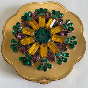 May include: A gold-toned compact with a floral design. The center features a large green jewel surrounded by yellow, purple, and green rectangular and oval-shaped jewels. The compact has a scalloped edge.