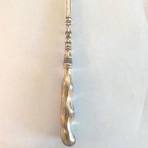 May include: A silver-toned crochet hook with a curved hook end and a decorative handle. The handle has a bulbous, ergonomic design for comfortable use. The hook is designed for yarn crafts.
