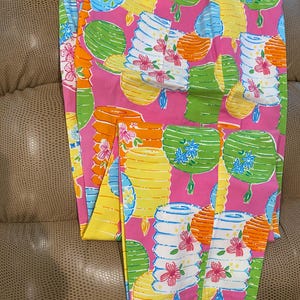 May include: Pink pants with a vibrant pattern of colorful lanterns in shades of yellow, orange, green, and blue. The lanterns are adorned with floral accents. The pants are folded, showcasing the full design.