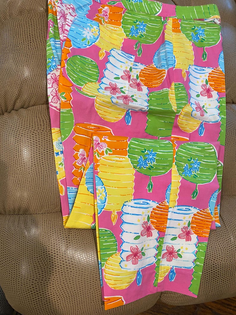 May include: Pink pants with a vibrant pattern of colorful lanterns in shades of yellow, orange, green, and blue. The lanterns are adorned with floral accents. The pants are folded, showcasing the full design.