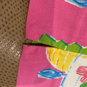 May include: Close-up of a pink fabric with a colorful floral design. The design includes a white background with blue, yellow, and pink accents. The fabric is trimmed with green and white edges.