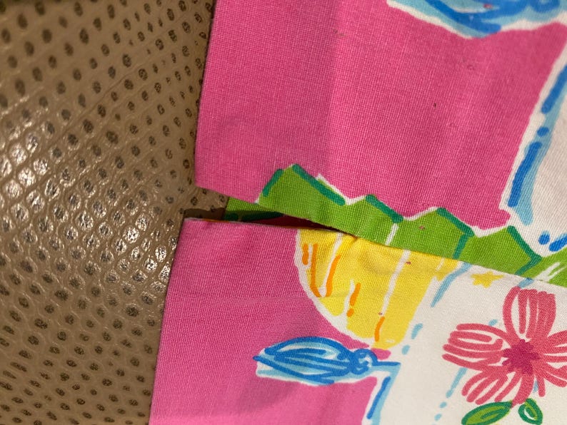 May include: Close-up of a pink fabric with a colorful floral design. The design includes a white background with blue, yellow, and pink accents. The fabric is trimmed with green and white edges.