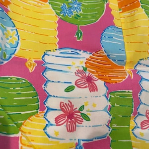 May include: A vibrant pink fabric featuring a repeating pattern of colorful lanterns. The lanterns are in shades of yellow, orange, green, and blue, with floral accents. The design is reminiscent of a festive, tropical theme.