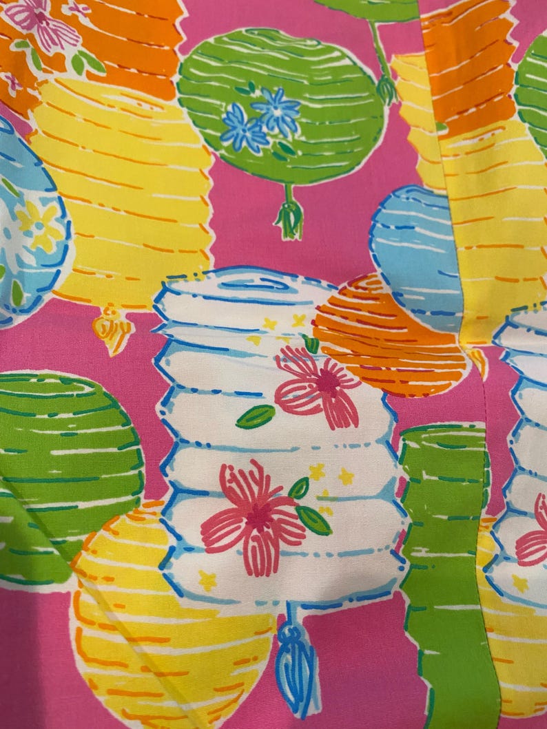 May include: A vibrant pink fabric featuring a repeating pattern of colorful lanterns. The lanterns are in shades of yellow, orange, green, and blue, with floral accents. The design is reminiscent of a festive, tropical theme.