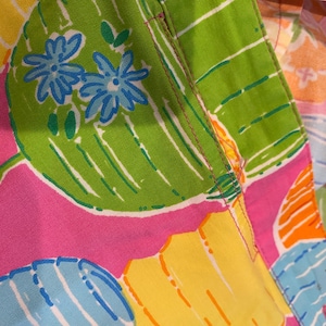 May include: Close-up of a colorful fabric with a vibrant floral pattern. The design features large green leaves with blue flowers, set against a pink, yellow, and blue background. The fabric has a stitched seam.