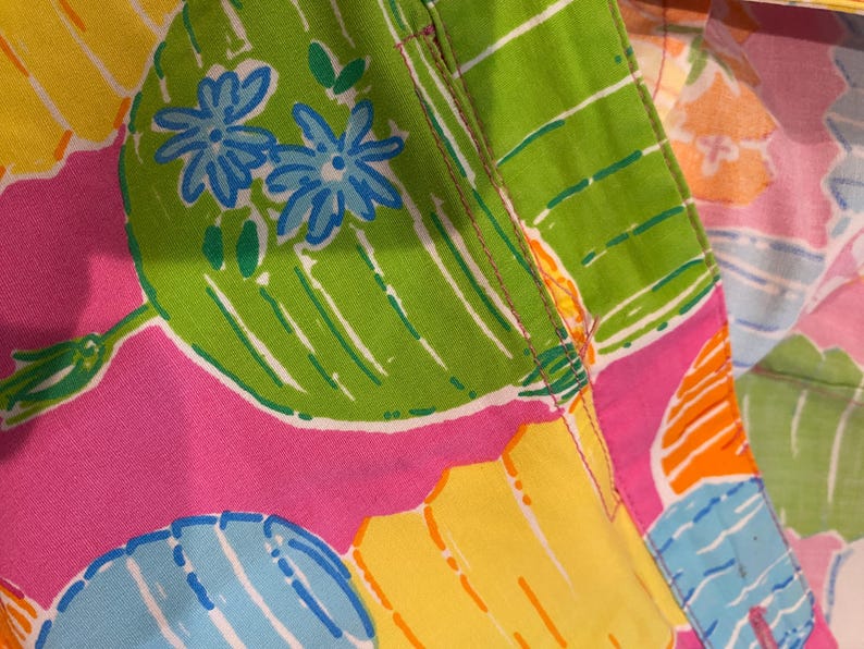 May include: Close-up of a colorful fabric with a vibrant floral pattern. The design features large green leaves with blue flowers, set against a pink, yellow, and blue background. The fabric has a stitched seam.
