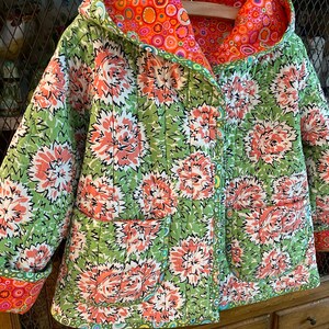 May include: A green quilted jacket with a floral print and a contrasting orange and pink lining. The jacket has a hood and two pockets.