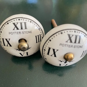 May include: Two white ceramic cabinet knobs with a clock face design. The knobs feature black Roman numerals and the text "POTTER-STONE". The hardware has a brass finish.