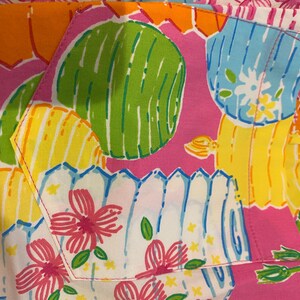 May include: Close-up of a colorful fabric with a repeating pattern of stylized flowers, cacti, and fence-like shapes in pink, green, yellow, blue, and orange. The fabric has a playful and vibrant aesthetic.