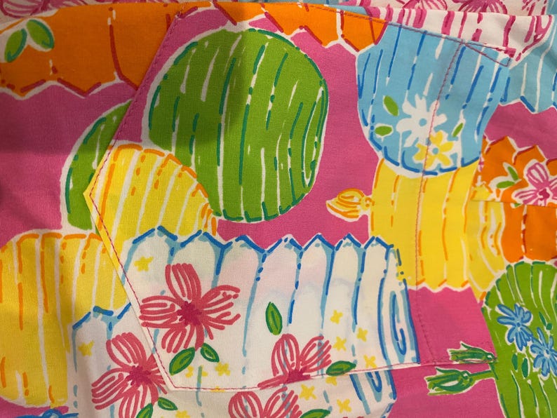 May include: Close-up of a colorful fabric with a repeating pattern of stylized flowers, cacti, and fence-like shapes in pink, green, yellow, blue, and orange. The fabric has a playful and vibrant aesthetic.