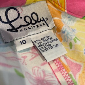 May include: Close-up of a colorful Lilly Pulitzer garment tag. The tag is white with black text, including the brand name and size 10. The fabric is a vibrant mix of pink, yellow, and floral patterns. The tag indicates the garment is made of cotton and spandex.