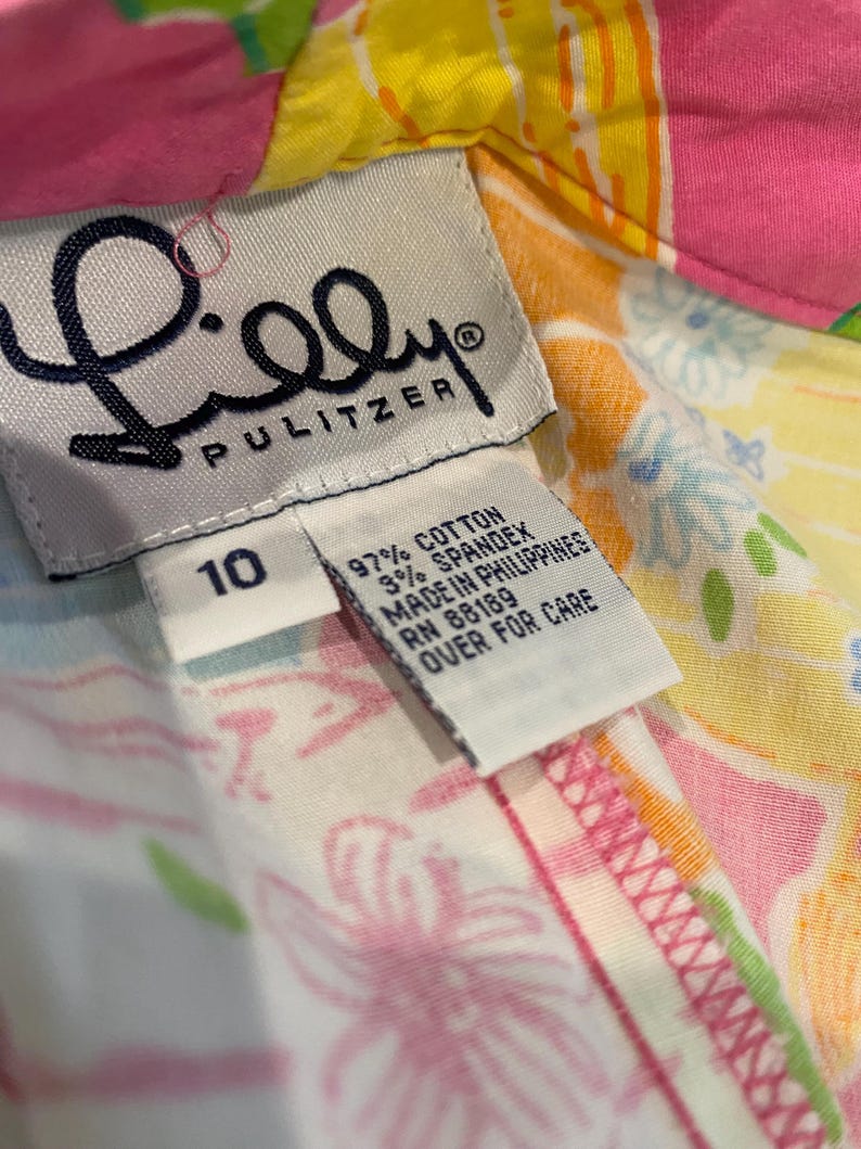 May include: Close-up of a colorful Lilly Pulitzer garment tag. The tag is white with black text, including the brand name and size 10. The fabric is a vibrant mix of pink, yellow, and floral patterns. The tag indicates the garment is made of cotton and spandex.