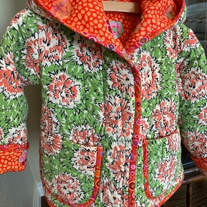 May include: A quilted jacket with a floral pattern in green, white, and coral. The jacket has a hood and two pockets. The interior and trim are a solid orange with a dotted pattern. The jacket is hanging on a wooden hanger.