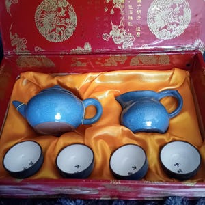 May include: A blue ceramic tea set comprising a teapot, creamer jug, and four small cups. The set is presented in a red box with gold detailing and a yellow satin lining. The teapot and jug have a crackle glaze finish.