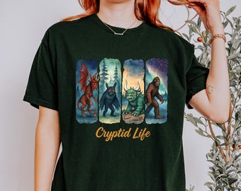 Può includere: T-shirt verde scuro con una grafica colorata di criptidi, tra cui un drago, una creatura nera, un mostro verde e Bigfoot. Il testo "Cryptid Life" è stampato sotto.