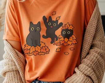 May include: An orange short-sleeved t-shirt featuring a graphic illustration of three black cats playing in piles of orange and brown autumn leaves. The cats have large yellow eyes and are depicted in various poses, including sitting and jumping.