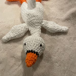 May include: A white crocheted goose plush toy with orange feet and beak lying on a white fuzzy surface.