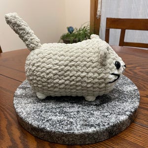 May include: A crocheted plush toy shaped like a cat, with a white and gray color scheme. The cat is sitting on a round gray stone base.