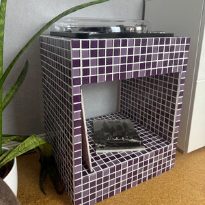 Op de afbeelding: Een paarse betegelde draaitafelstandaard met een plank om platen op te bergen. De standaard heeft een transparante draaitafel bovenop en een boek met de titel "New York" op de plank.