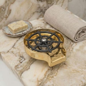 May include: A gold-colored soap dispenser with a black, ship's wheel-shaped top. A bar of soap sits in a small, decorative dish. A rolled-up, beige towel is also present. The items are on a marble countertop.