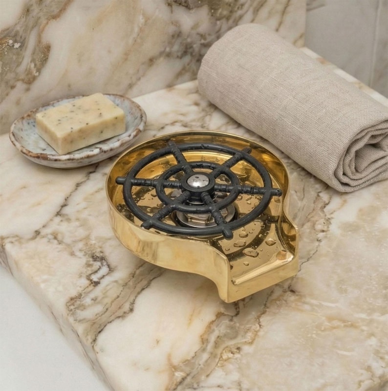 May include: A gold-colored soap dispenser with a black, ship's wheel-shaped top. A bar of soap sits in a small, decorative dish. A rolled-up, beige towel is also present. The items are on a marble countertop.